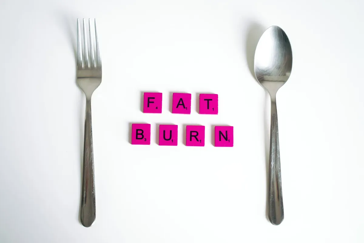 Conceptual art featuring the words 'Fat Burn' using Scrabble tiles between a fork and spoon on a white background.